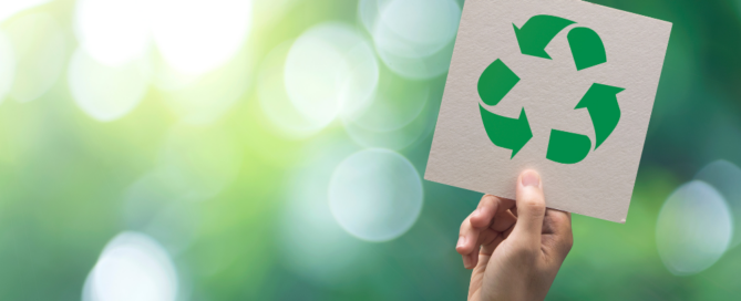 Photo shows a hand holding up a green recycling sign with greenery in the background